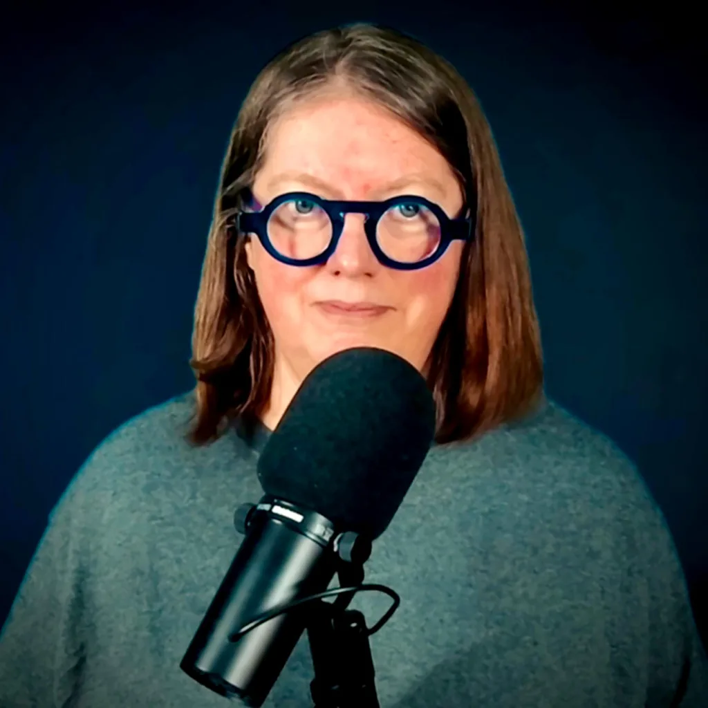 jen dehaan standing behind a microphone in grey tshirt and round blue glasses